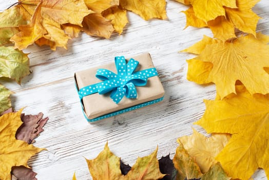 Flat lay autumn composition with gift box and yellow autumn leaves. Holiday present decorated blue ribbon bow on vintage wooden desk. Happy thanksgiving congratulation. Autumn sale advertising.