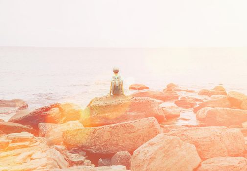 Traveler young woman with backpack resting on big stone near the sea in summer, rear view. Image with sunlight effect