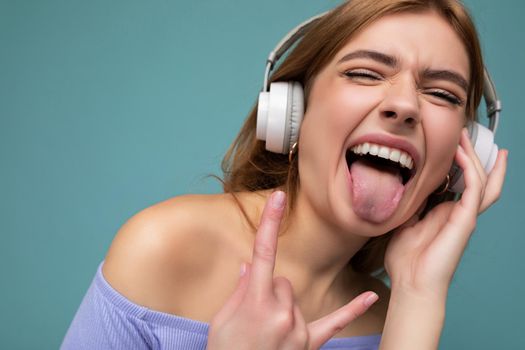 Closeup portrait of attractive emotional young blonde woman wearing blue crop top isolated on blue background wearing white wireless bluetooth headphones listening to cool music and having fun showing tongue and rock and roll gesture.