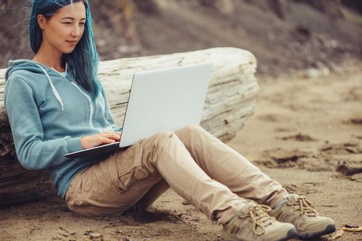 Smiling freelancer girl with blue hair working on laptop on sand beach. Freelance concept