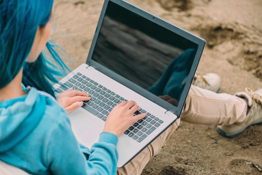 Freelancer girl with blue hair working on laptop on sand beach. Freelance concept. Space for text on screen of laptop