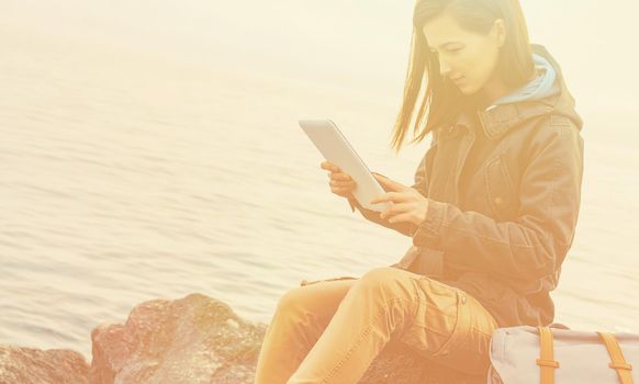 Traveler girl sitting on coast near the sea and working on digital tablet. Image with sunlight effect