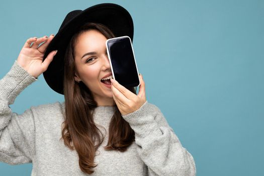 closeup Photo of Beautiful positive woman person wearing black hat and grey sweater holding mobilephone showing smartphone isolated on background looking at camera.Mock up, cutout, free space