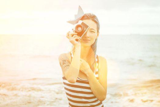 Beautiful young woman in costume of sailor takes a photograph with old photo camera on beach in summer. Image with sunlight effect