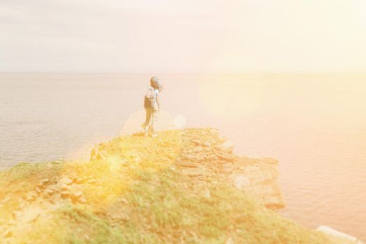 Hiker young woman with backpack standing on peak of mountain and enjoying view of sea in summer. Image with sunlight effect