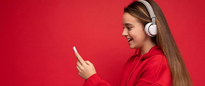 Panoramic Side-profile Shot of positive smiling pretty brunette girl wearing red hoodie isolated on red background holding and using smartphone writing sms message wearing white wireless headphones listening to cool music looking at phone display.Copy space