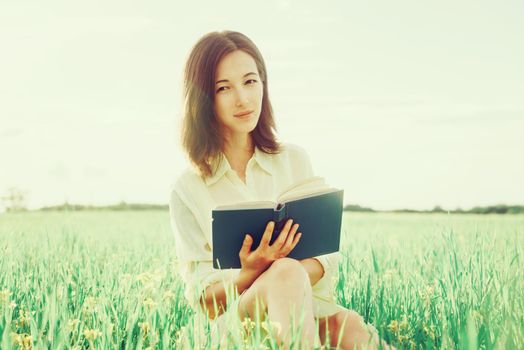 Beautiful young woman resting with book on summer flower meadow, woman looking at camera