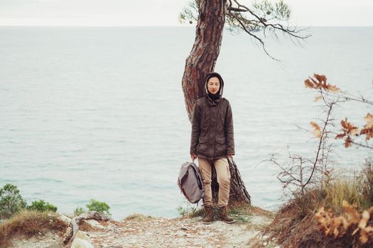 Traveler young woman wearing in warm clothing with backpack standing near a tree on background of sea and looking at camera