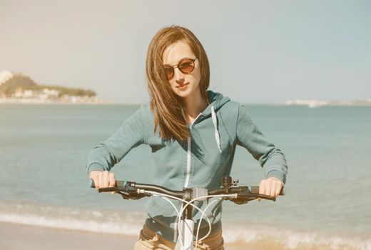 Smiling young woman in sunglasses with bicycle on beach on background of sea in summer at sunny day
