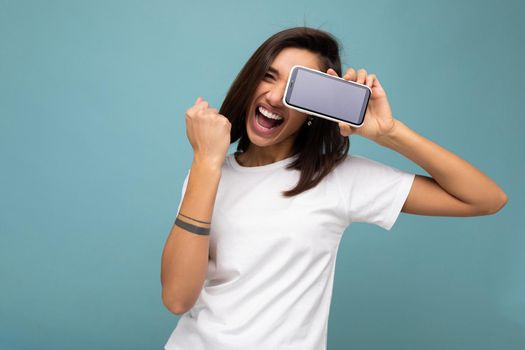 Attractive smiling young brunet woman good looking wearing white t-shirt standing isolated on blue background with copy space holding smartphone showing phone in hand with empty screen screen for cutout showing yes gesture.