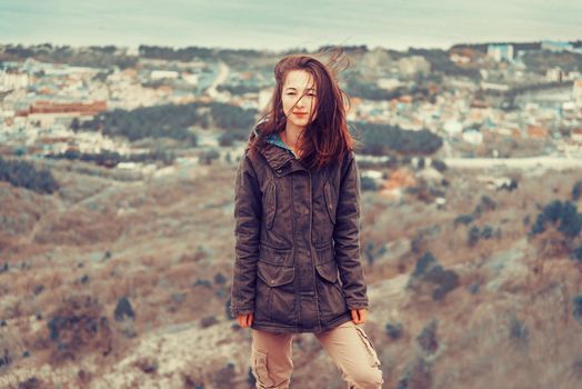 Young woman dressed in parka jacket and cargo pants standing on hill on background of town
