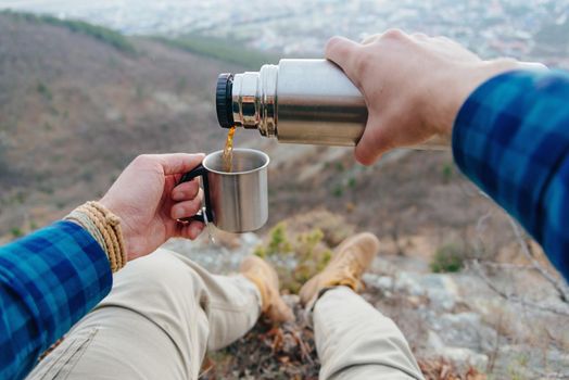 Hiker man sitting in mountains and pouring tea from thermos to cup. Point of view shot