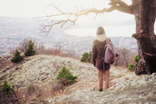 Hiker young woman standing on peak of hill and looking at the town, rear view