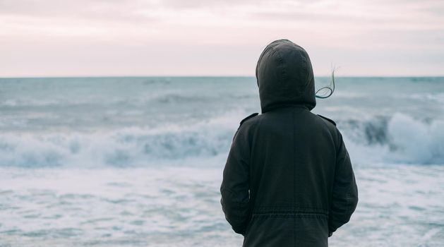 Unrecognizable traveler young woman in hood looking at sea waves in windy weather, rear view