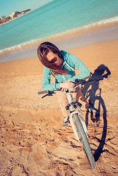 Girl in sunglasses with bicycle standing on sand beach near the sea in summer