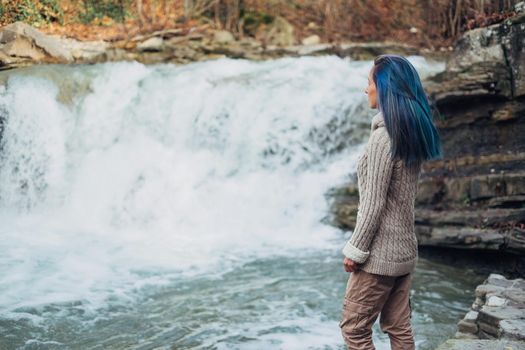 Beautiful young woman with blue hair standing near the waterfall