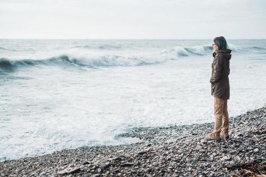 Traveler young woman standing on pebble coast and looking at sea waves in windy weather