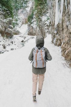 Hiker young woman with backpack walking on snowy footpath near the cliff, rear view