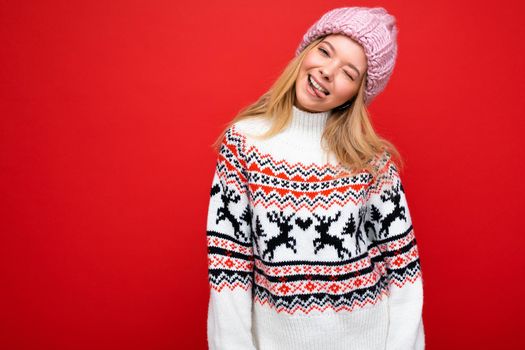 Photo of pretty positive happy joyful young blonde female person isolated over red background wall wearing winter sweater and pink hat looking at camera winking and showing tongue. Copy space