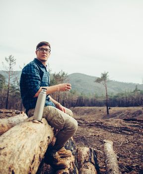 Young man sitting on fallen tree trunk and drinking tea from thermos. Handsome man resting outdoor