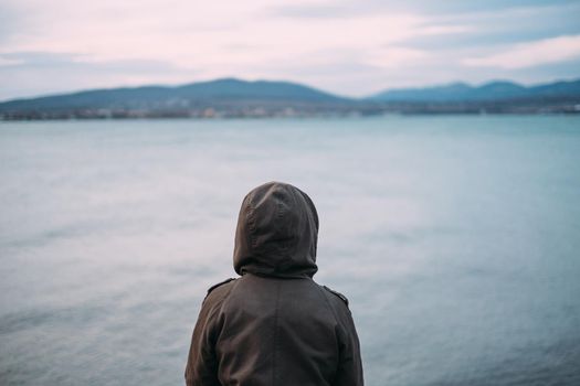 Unrecognizable woman wearing in parka jacket with hood enjoying view of mountain and sea bay in evening, rear view