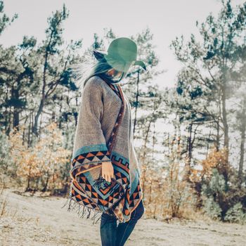 Fashionable young woman in hat and poncho walking in forest among pine trees