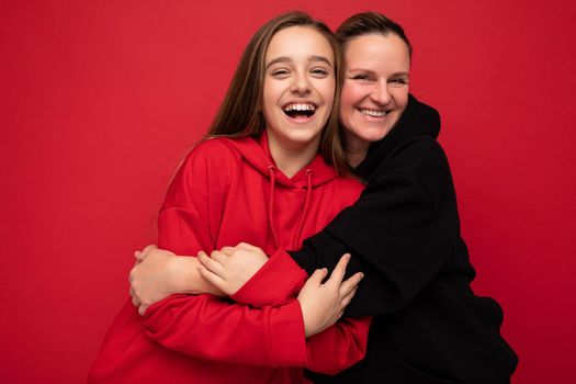 Photo shot of pretty positive smiling brunette female teenager wearing stylish red hoodie and adult brunette woman wearing trendy black hoodie standing isolated over red background wall looking at camera and having fun.