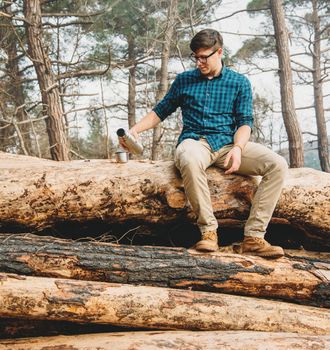 Traveler young man sitting on tree trunk and pouring tea from thermos to cup in the forest. Man has a picnic outdoor