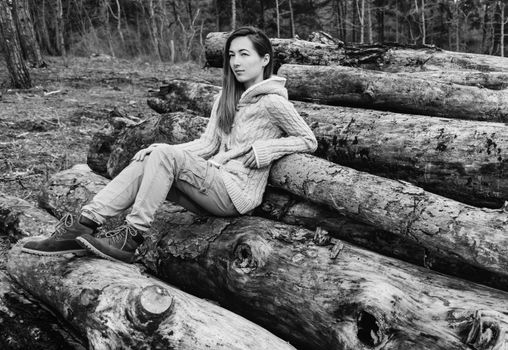 Young woman sitting on stack of tree trunk, girl resting outdoor. Black and white image