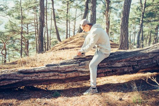 Sporty young man sitting on fallen tree trunk and tying shoelaces outdoor in the pine forest