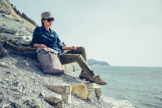Traveler young woman sitting on stone coast with backpack and enjoying view of sea in summer