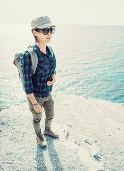 Smiling beautiful hiker girl in sunglasses with backpack standing on steep coast near the sea in summer outdoor