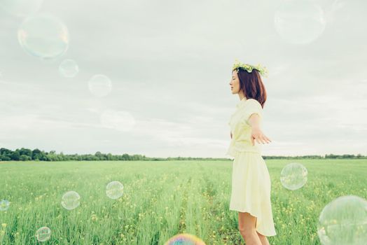 Happy beautiful young woman in flower wreath walking on summer meadow among soap bubbles