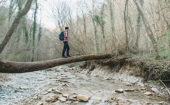 Hiker young man with backpack crossing river on fallen tree trunk in the forest