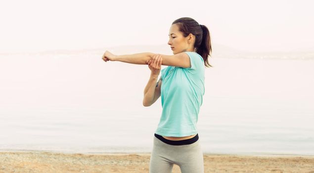 Sporty young woman stretching her arms on beach in summer in the morning, workout. Concept of healthy lifestyle