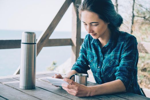 Tourist young woman sitting in summer wooden veranda outdoor with thermos and mobile phone