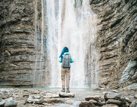 Traveler young woman with backpack standing on stone and enjoying view of beautiful waterfall outdoor, rear view