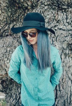 Beautiful young woman with blue hair wearing in hat and sunglasses standing on background of tree trunk