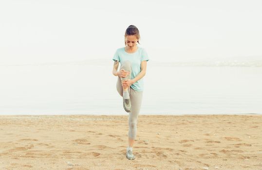 Sporty young woman stretching her legs and preparing to run on sand beach in summer, workout. Concept of healthy lifestyle