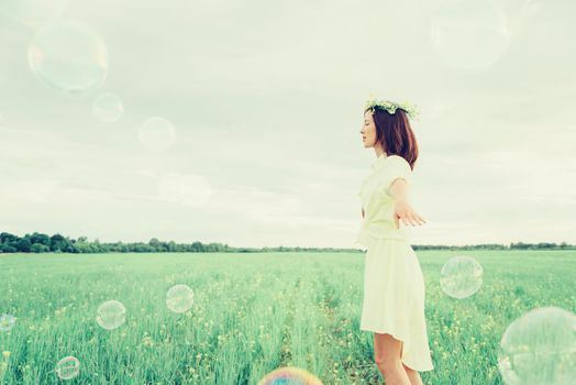 Happy beautiful girl in flower wreath walking on summer meadow among soap bubbles