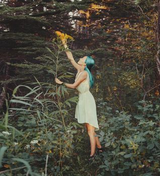 Beautiful girl with blue hair looking at big yellow flower in summer forest