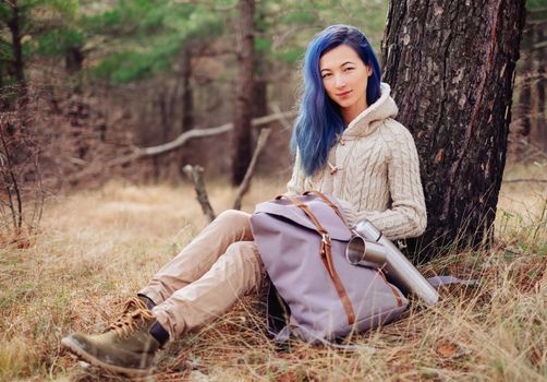 Beautiful young woman resting near a tree in the forest with backpack, looking at camera