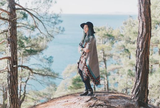 Beautiful young woman wearing in poncho and hat standing among pine trees in the summer forest on background of sea