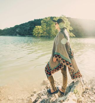 Beautiful fashionable young woman wearing in hat and poncho standing on coast and looking at lake in summer