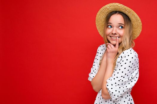 Young beautiful woman. Trendy woman in casual summer dress and straw hat. Positive female shows facial emotions. Funny model isolated on red background with free space.