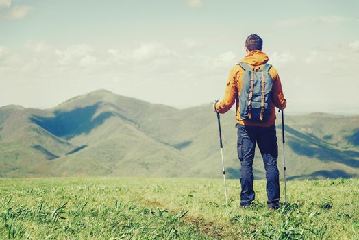 Hiker young man with backpack and trekking poles looking at mountains in summer outdoor. Free space in left part of the image