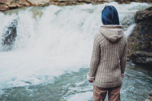 Traveler young woman with blue hair looking at beautiful waterfall, rear view