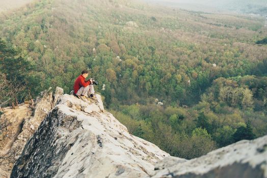 Traveler photographer young man sitting on peak of rock and take a photograph of landscape in the summer mountains