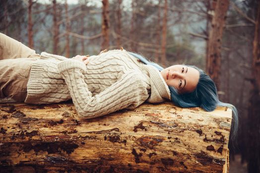 Beautiful young woman with blue hair lying on fallen tree trunk and looking at camera. Smiling girl resting outdoor