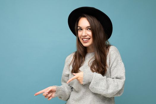 Photo of pretty brunette positive young woman directing fingers to the side with empty space, demonstrating presenting low prices poster, wearing grey sweater and black hat, isolated on blue background.